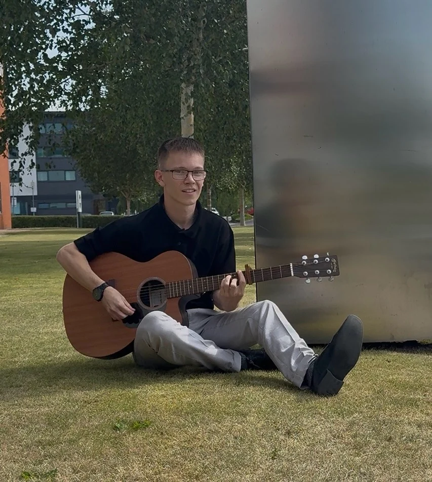 Harry Basnett sitting outdoors with his acoustic guitar
